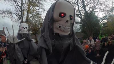 Two lanky skeletons with black capes on are towering above a crowd of people and in various dancing poses. Behind is Lincoln Cathedral in the distance.