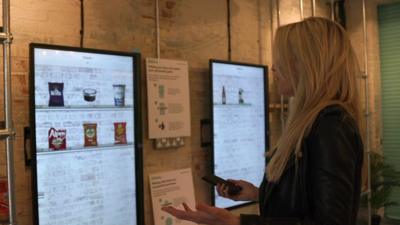 Lara Lewington stands in front of touch screens displaying a range of products