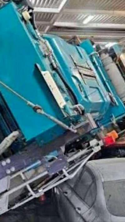 A large blue machine is on its side on a grey car and a white van. A worker in a hard hat and overalls inspects the scene.