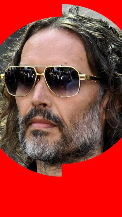 Russell Brand wearing sunglasses - he has long curly brown hair and a beard  