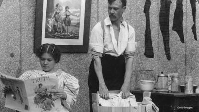 A woman reading a magazine while a man washes the laundry