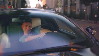 Alasdair Keane sits behind the wheel of a car, appearing to swerve