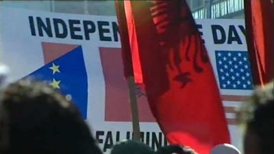 People wave the New Flag of Kosovo. A banner in the background says 'Independence Day'