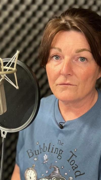 Gayanne sits in front of a microphone in a grey soundproofed recording booth, she has brown hair and wears a blue T-shirt