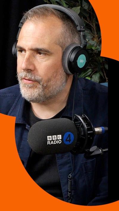 A man with short, grey hair sits in front of a microphone and looks at someone off-camera