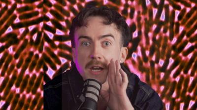 A man is holding his hand to his mouth and is speaking into a mic. He has dark brown short hair, blue eyes and a light brown moustache. He is wearing a dark denim jacket.The background is a close-up image of germs.