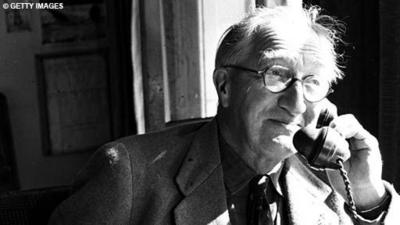 Black and white image of Sir Alan P Herbert on the telephone with a slight grin on his face.