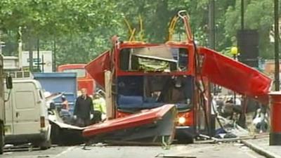 A red London bus with metal and debris protruding from what was once the roof, with emergency workers nearby.