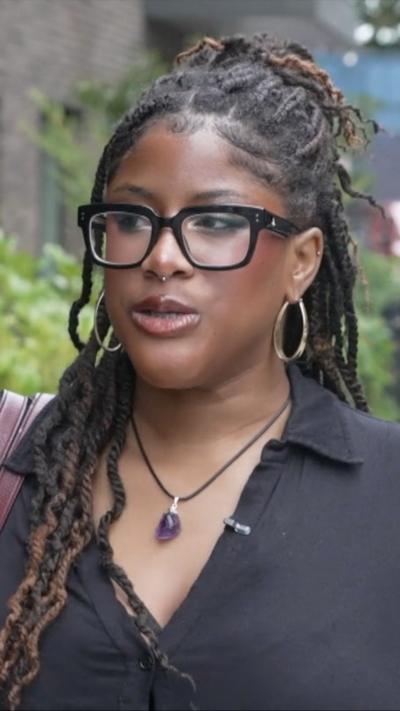 Woman with dreadlocks, black shirt, and purple necklace standing on a sidewalk near a brick building and greenery.