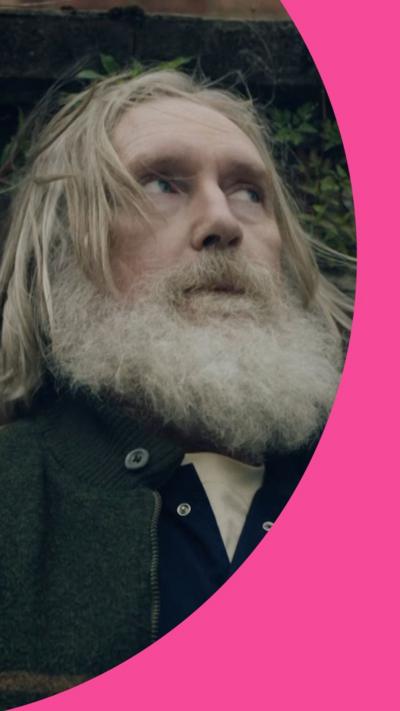 A man with long grey hair and a beard stands by a wall looking to the side.