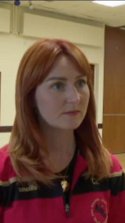 A redheaded woman in an athletic zip-up jumper, looking at somebody outside the frame. Her facial expression is a little sad or worried