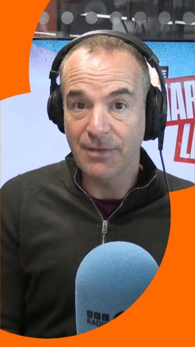 Martin Lewis looks straight at the camera. He wears black headphones and sits infant of a blue microphone.