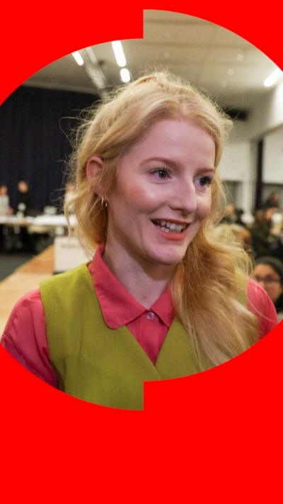 Hannah Spencer has blonde hair and is wearing green top over a pink shirt