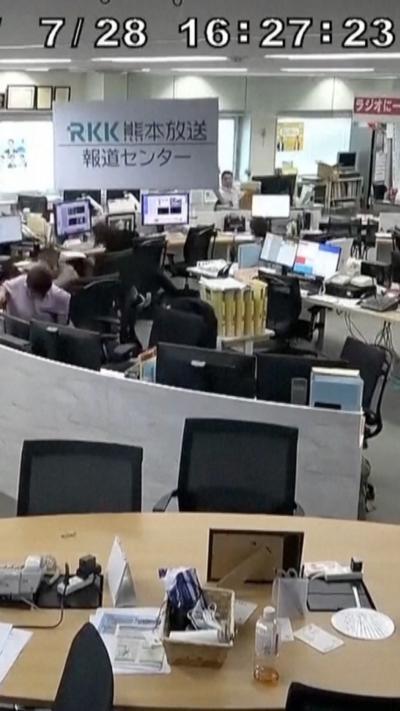 A man in a shirt ducks beneath a desk, surrounded by shaking office furniture and screens.