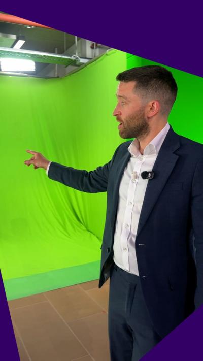 Political Correspondent David Wallace Lockhart looks off camera as he points to a green screen inside the BBC Scotland building. He has short brown hair, brown stubble and is wearing a white shite and dark grey suit.