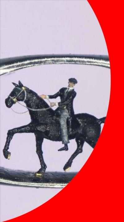 A micro sculpture of a man in a black coat, black hat and grey trousers sat in a horse inside the eye of a needle