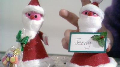 Two Santa Christmas decorations made out of card, tissue paper and sticky-backed felt, one holding a namecard with 'Jenny' written on it.