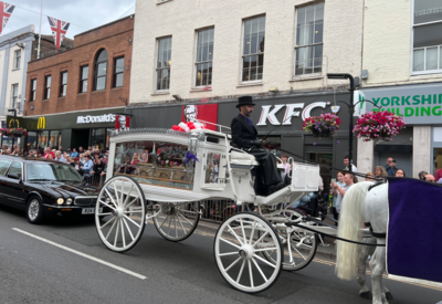 A horse is pulling a white carriage with Pauline's coffin inside it. On the pavement are many people lining up to pay their respects, and stood outside the KFC she worked at before she died.