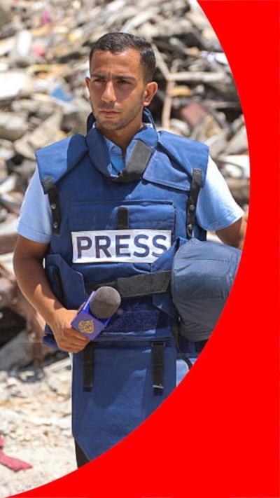 Anas al-Shariff, wearing a blue Press vest and carrying helmet and mic, stands in front of a pile of rubble, 2024