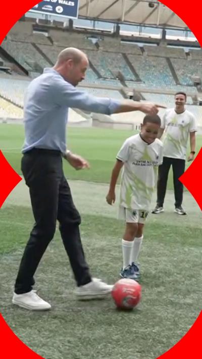 William standing in stadium with red ball at his feet. He is pointing forward and there's a young boy in a white and green football kit smiling next to him.