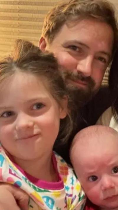 Man with a beard smiling and holding a girl in a colourful top and baby boy