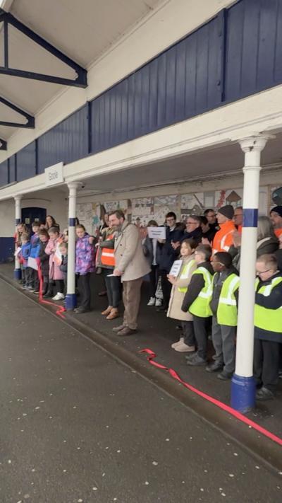 A group of children and adults are gathered on a train station platform unveiling the mural. Some people are taking photos while others clap, with a colourful mural providing a bright backdrop behind the crowd.