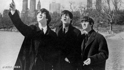 Black and white. John Lennon, Paul McCartyney and Ringo Starr with the New York skyline behind them, looking off in the distance.  Ringo smoking, John pointing.