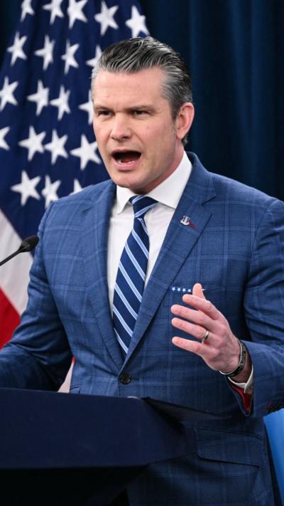 Hegseth speaks into a microphone standing in front of an American flag wearing a blue suit.