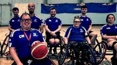 A wheelchair basketball team wearing blue sports tops and black shorts are smiling at the camera. Four men are on the back row and two women and one man are on the front row. 
