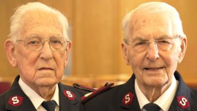 Maurice and George, both in their 90s who have been friends for 87 years. They are both older men with white hair and both are wearing a black and red branded Salvation Army uniform