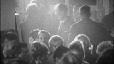 Black and white image of clubgoers at Greasbrough working man's club.  Some are sat down and some are standing up behind.  There is low level light and a beam of light shining through the middle of the image.