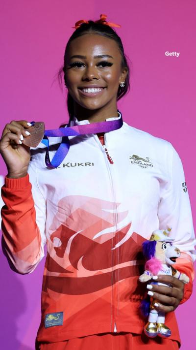 Shantae-Eve Amankwaah with her medal. She is holding it away from her neck and smiling. In her other hand is a small stuffed toy.
