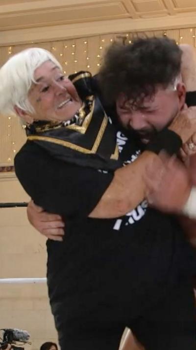 A nan with White hair and a black t-shirt and black and gold bandana putting a man in a headlock.