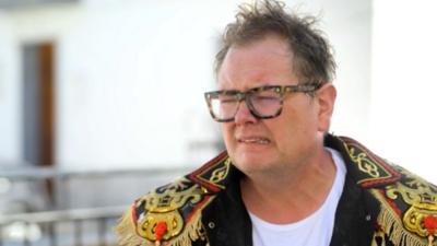 Alan Carr cringing, wearing glasses and a Spanish jacket.