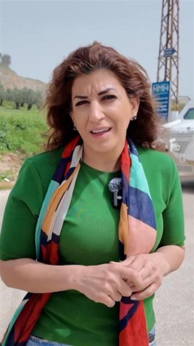 Carine Torbey, a woman with long brown hair, holds her hands in front of her chest, wearing a milt-coloured scarf and green top. with a white armoured vehicle behind her
