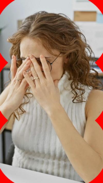 A woman with long brown hair, sat at a desk, covering her face with her hands.