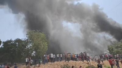 A group of people stand near a plane crash site, as black smoke billows into the air.