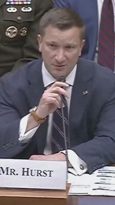 A man holds a microphone with his right hand while wearing a blue suit and white shirt.