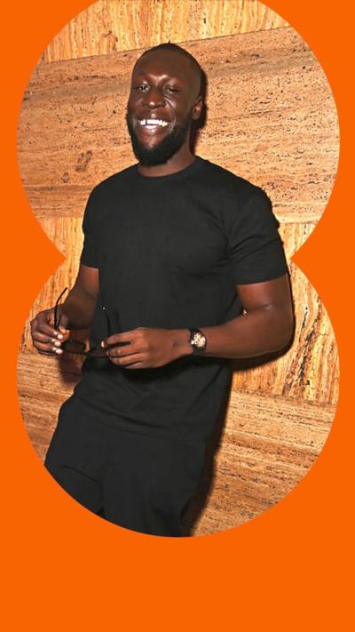 Stormzy leans onto a wall whilst smiling and holding up a pair of sunglasses