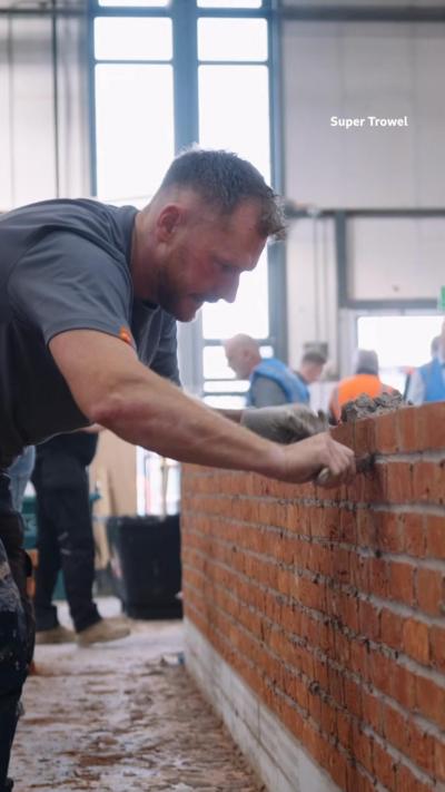 A man in a grey t-shirt smoothing cement on a red brick wall. 