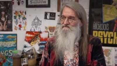 Rob Davies in his shop. He is sitting in front of a wall decorated with eclectic, colourful film posters. He is looking away from the camera. He has long white hair and a long white beard. He is wearing round-rimmed glasses.