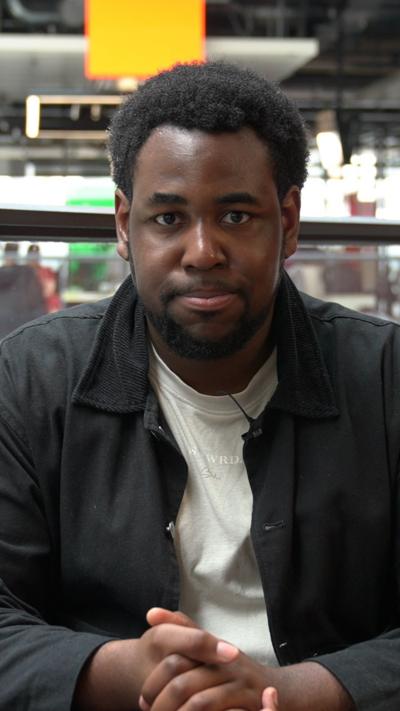 A man with a black jacket and a cream t shirt is looking directly into the camera. He is sat on an orange sofa, with glass barriers, desks and windows behind them. 
