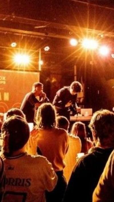 Two performers with a DJ deck on stage, with a crowd of people in front. One performer is holding a mic, the other is working on the DJ deck. Bright lights are above the stage and give the room and yellow-orange glow.