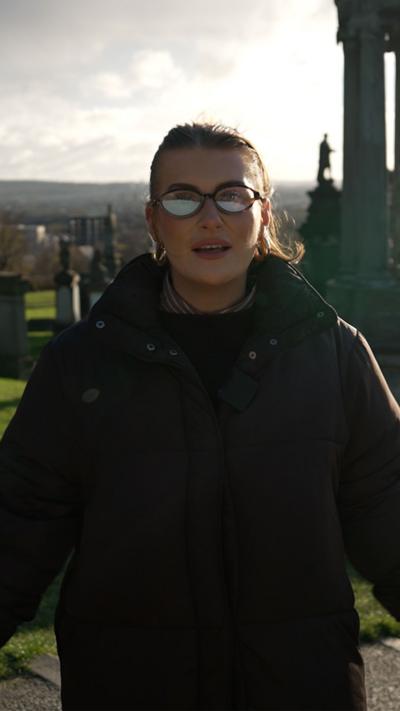 Woman in graveyard with glasses and brown jacket