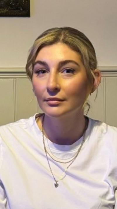 A woman with blonde hair tied up looking at the camera. She has a white t-shirt on. 