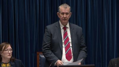 Edwin Poots is wearing a grey suit with a white shirt and a red tie with white and blue slanted stripes. He has short grey hair.