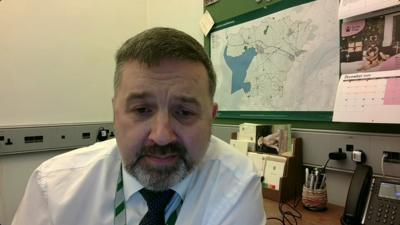 Robin Swann sitting an office desk. He has short brown hair and greyish beard, wearing a white shirt and red tie.