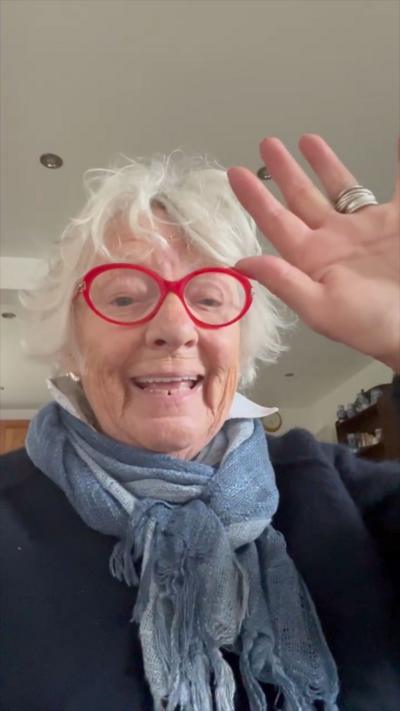 White haired Viv Truran, wearing red rimmed glasses and a blue scarf and jumper, smiles and waves to the camera