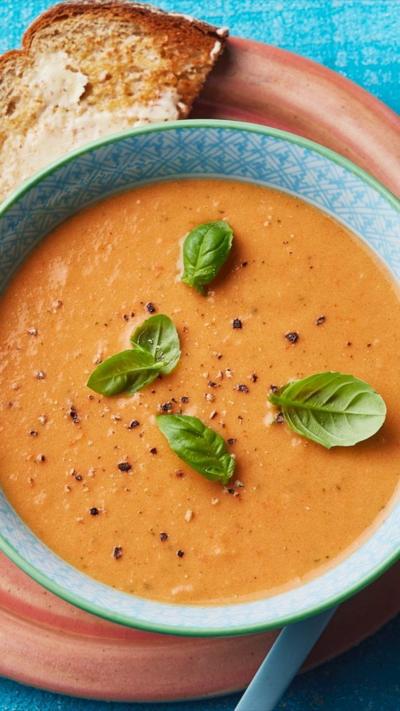 A blue bowl filled with tomato soup. Basil leaves are sprinkled on top. It's on a red plate with a piece of buttered toast.