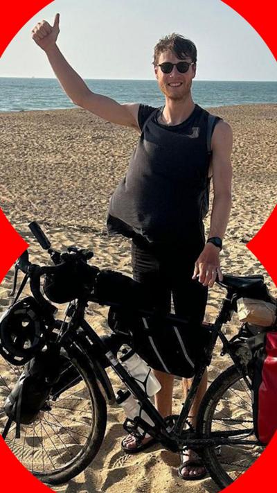 Man standing on a beach next to his bike with his arm in the air giving a thumbs up
